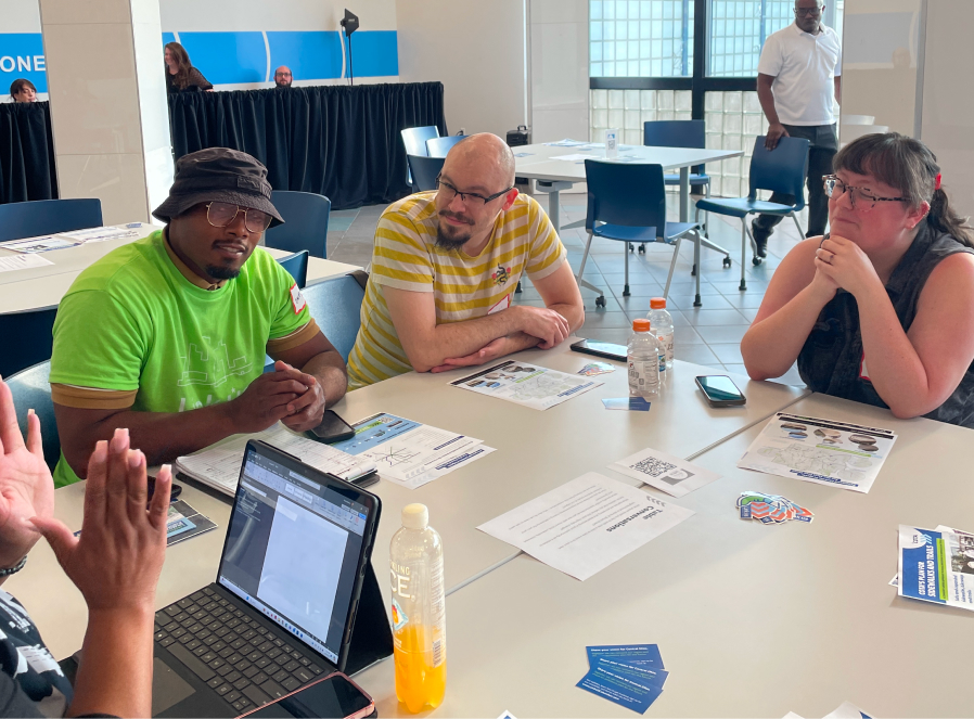 Two men and a women sit at a table looking at a speaker who is not shown, an open laptop and LinkUS printouts sit on the table.