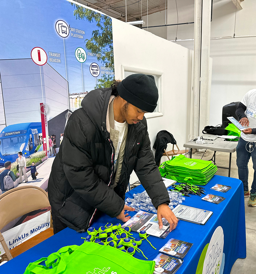 Person at an indoor event table setting up green branded bags and materials, with a transit route map displayed behind them.