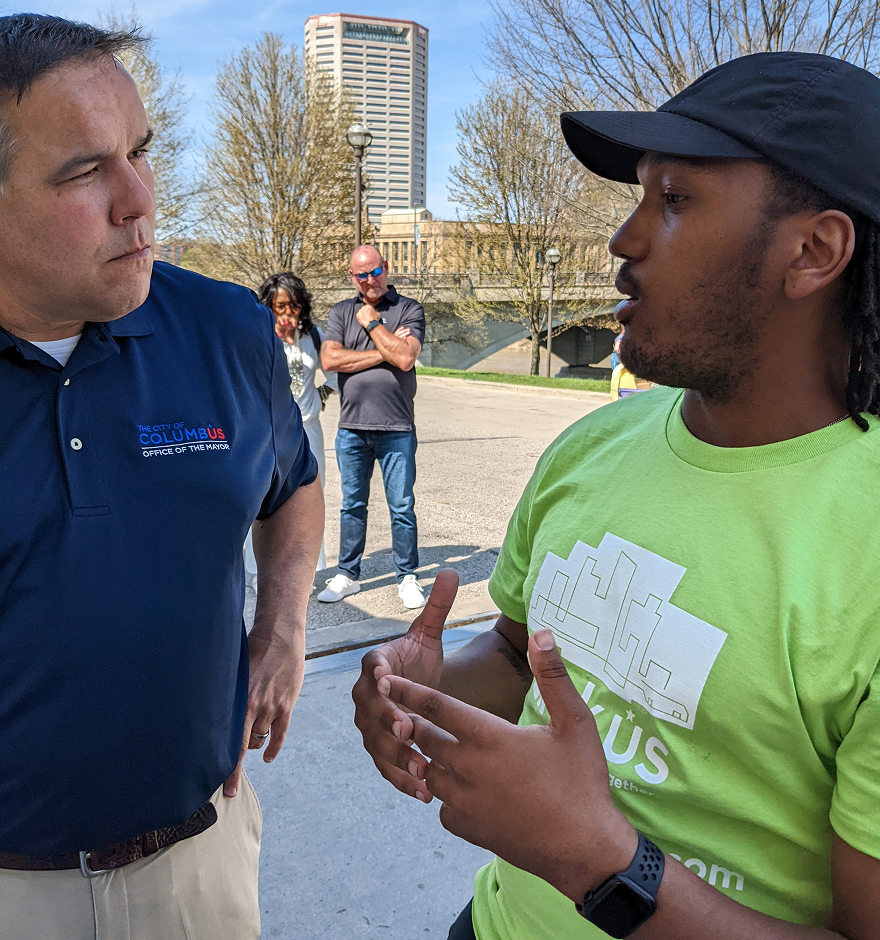 Two men in conversation outdoors along the riverfront, one wearing a navy polo and the other a green shirt with a hand raised mid-gesture, with the Columbus skyline in the background. The man on the left is Mayor Andrew Ginther.