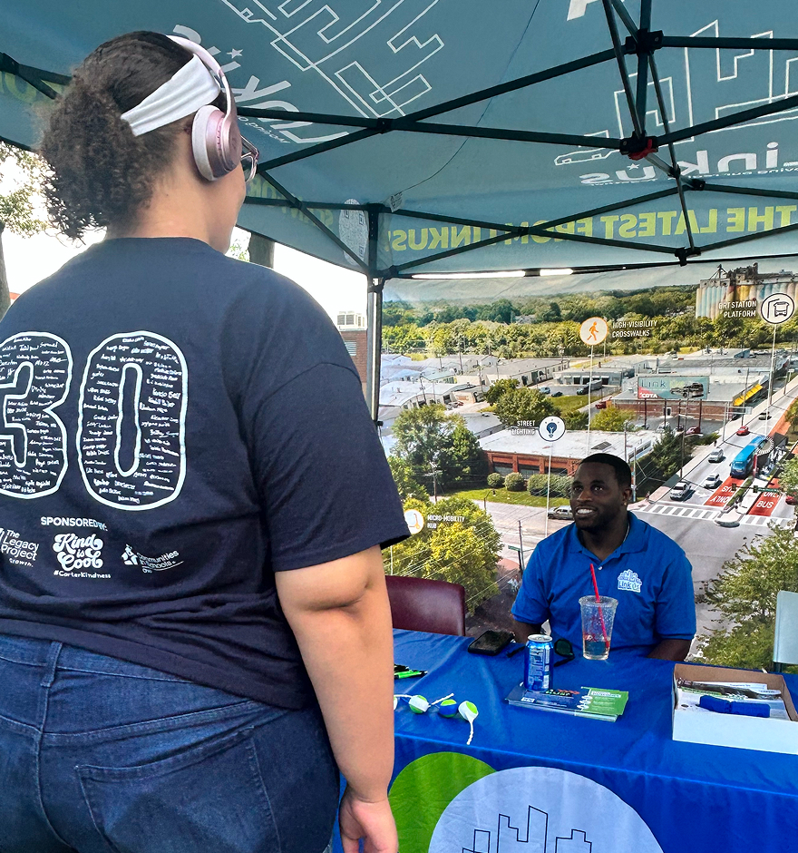 COTA staff member at an outdoor event tent booth, with a community member wearing a headband looking at branded materials on the table.